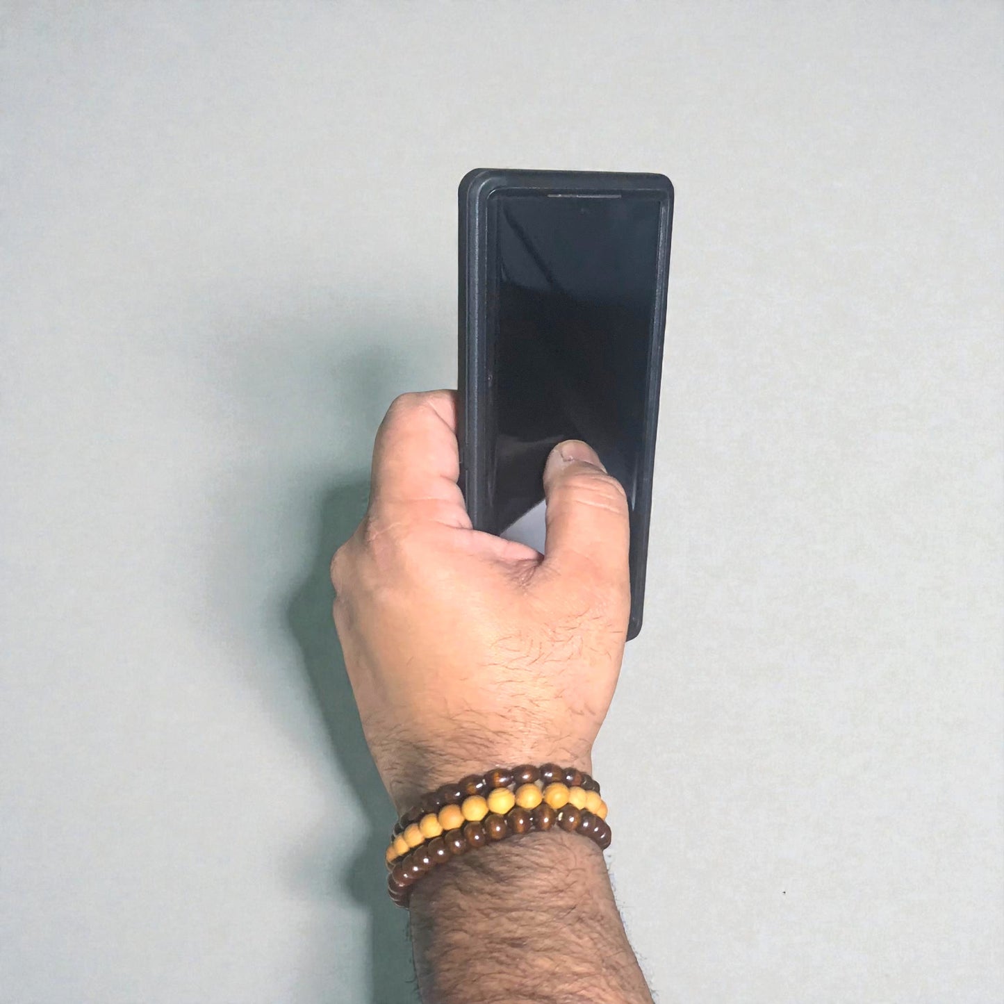 Hand holding a black smartphone against a light gray background. The focal point is a 3 stack of mixed colour wooden bracelets.