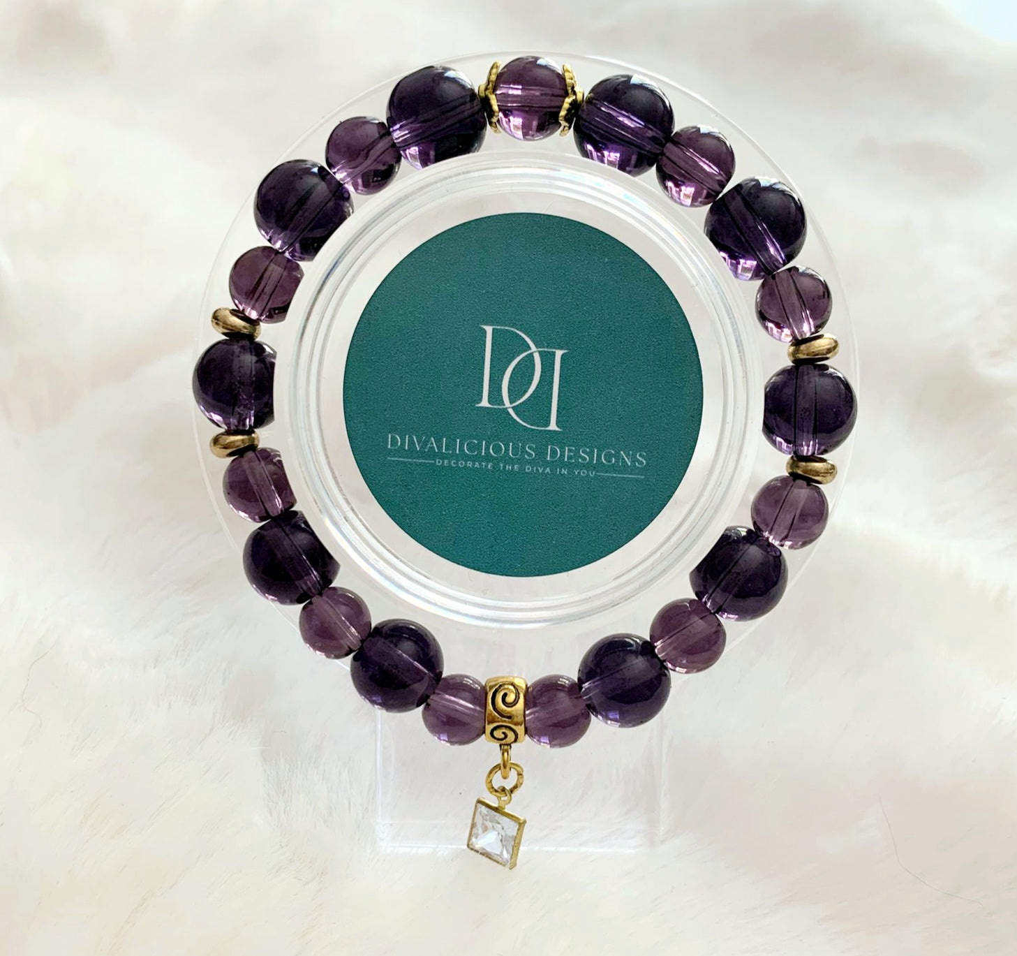 An amethyst glass bracelet with gold accents displayed in a clear case, featuring a square cubic zirconia charm.