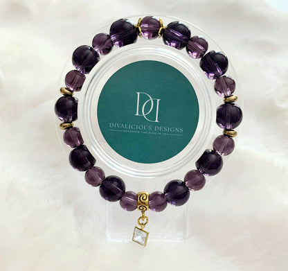 An amethyst glass bracelet with gold accents displayed in a clear case, featuring a square cubic zirconia charm.