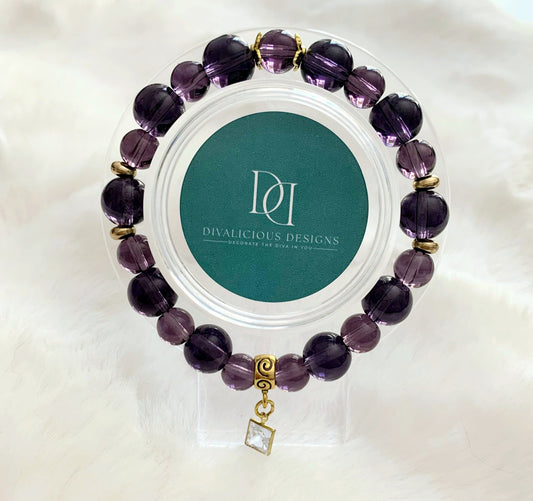 An amethyst glass bracelet with gold accents displayed in a clear case, featuring a square cubic zirconia charm.