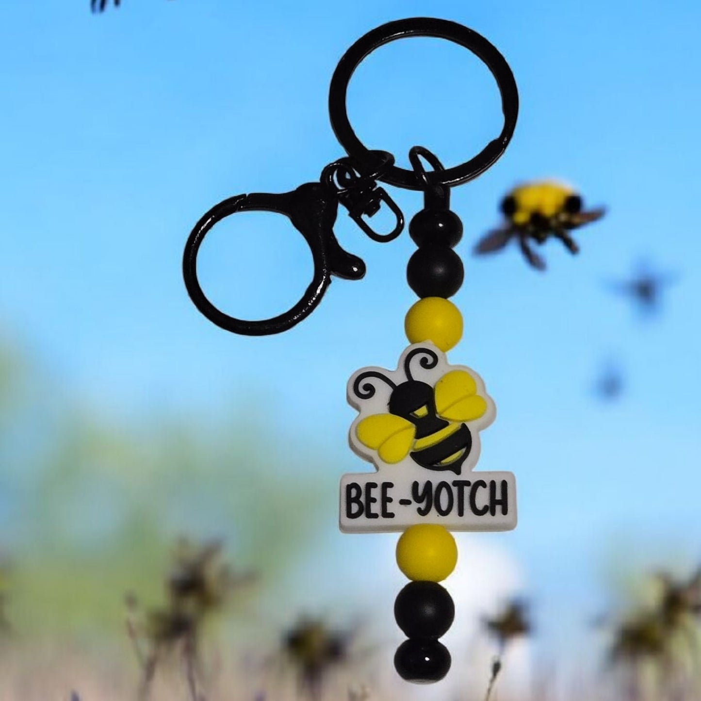 Keychain with bee design and 'BEE-YOTCH' text against a blurred natural background with flowers and bees.