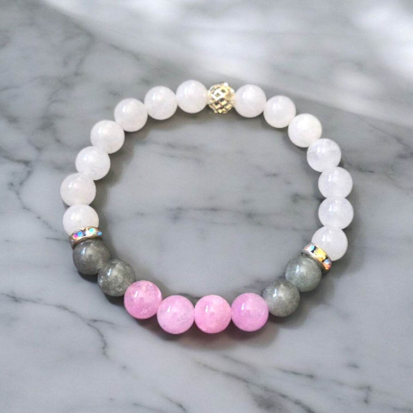 White chalcedony bracelet with pink kunzite, moss agate beads, rhinestone spacers, and a gold cage bead displayed on a marble surface.