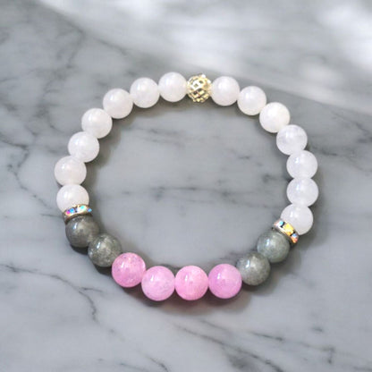White chalcedony bracelet with pink kunzite, moss agate beads, rhinestone spacers, and a gold cage bead displayed on a marble surface.