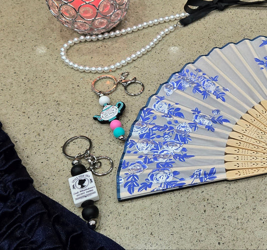 Decorative vintage folding fan with blue floral patterns, Bridgerto and Spilling the Tea keychains and accessories placed by candle on a textured surface.