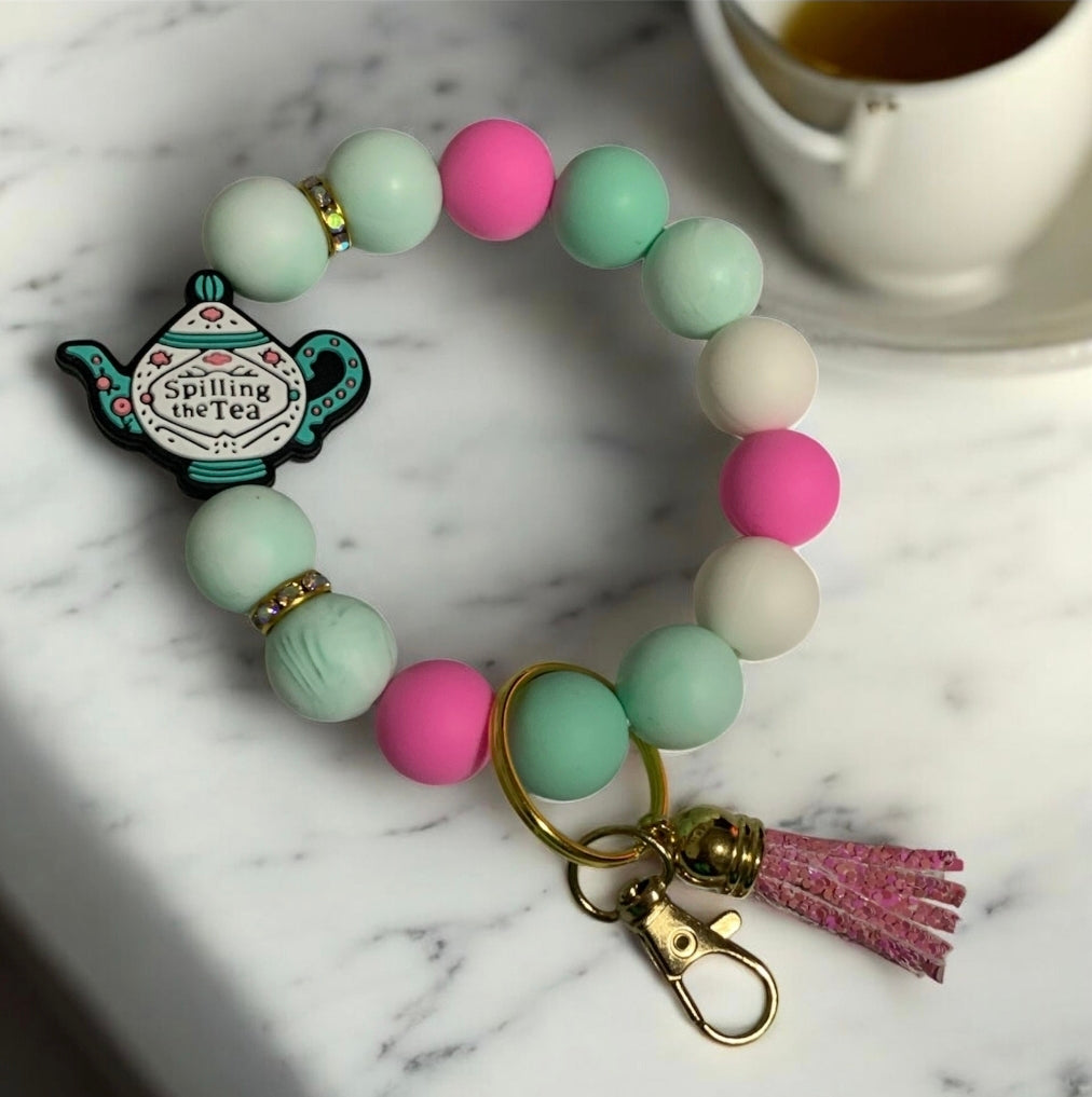 Silicone Keychain wristlet with a "Spilling the Tea" teapot charm on a marble surface with a cup of tea. Inspired by Bridgerton and Lady Whistledown