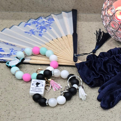 Decorative vintage folding fan with blue floral patterns, Bridgerto and Spilling the Tea keychain writslets and accessories placed by candle on a textured surface.