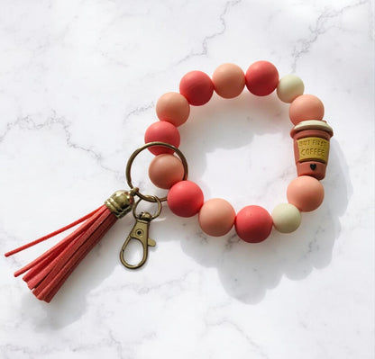Colorful beaded bracelet with a coffee cup charm and tassel on a marble background. The focal bead is a 3D coffee cup that says, "But First Coffee".