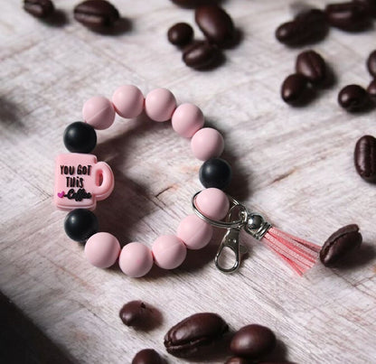 Pink beaded keychain bracelet with a focal bead saying,  "You Got This! - Coffee". The product is displayed on a wooden surface with coffee beans.