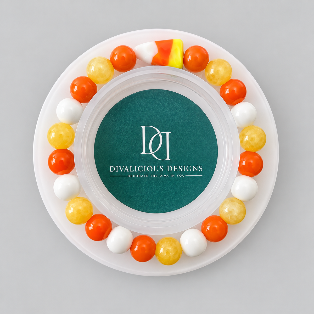 A round white tray or plate with a circular teal center printed with the brand name 'DIVALICIOUS DESIGNS' and white monogram lettering. Around the rim are alternating orange, yellow, and white candy-like gumballs, with a small candy corn piece at the top, all photographed from above on a light gray background.