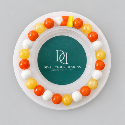 A round white tray or plate with a circular teal center printed with the brand name 'DIVALICIOUS DESIGNS' and white monogram lettering. Around the rim are alternating orange, yellow, and white candy-like gumballs, with a small candy corn piece at the top, all photographed from above on a light gray background.