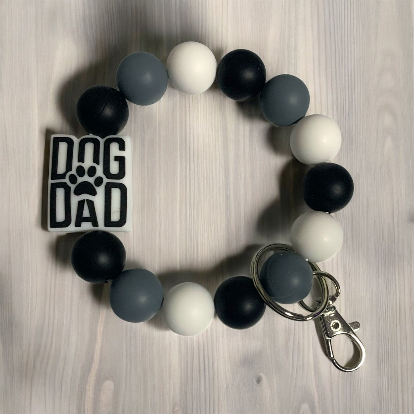 A silicone keychains bracelet with black, gray, and white beads and 'DOG DAD' focal bead displayed on a wooden surface.