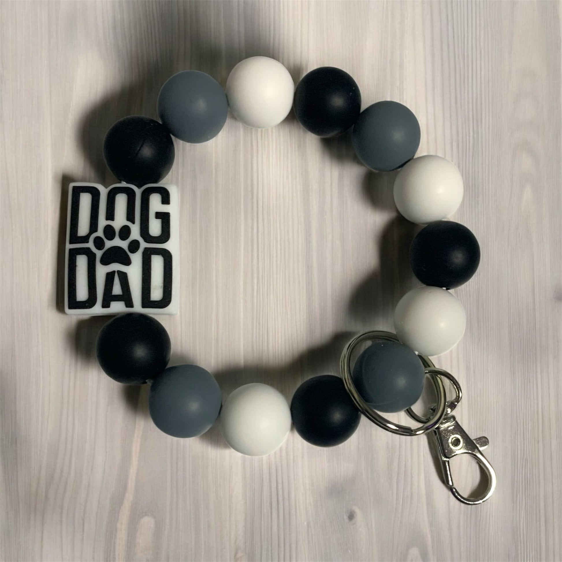 A silicone keychains bracelet with black, gray, and white beads and 'DOG DAD' focal bead displayed on a wooden surface.