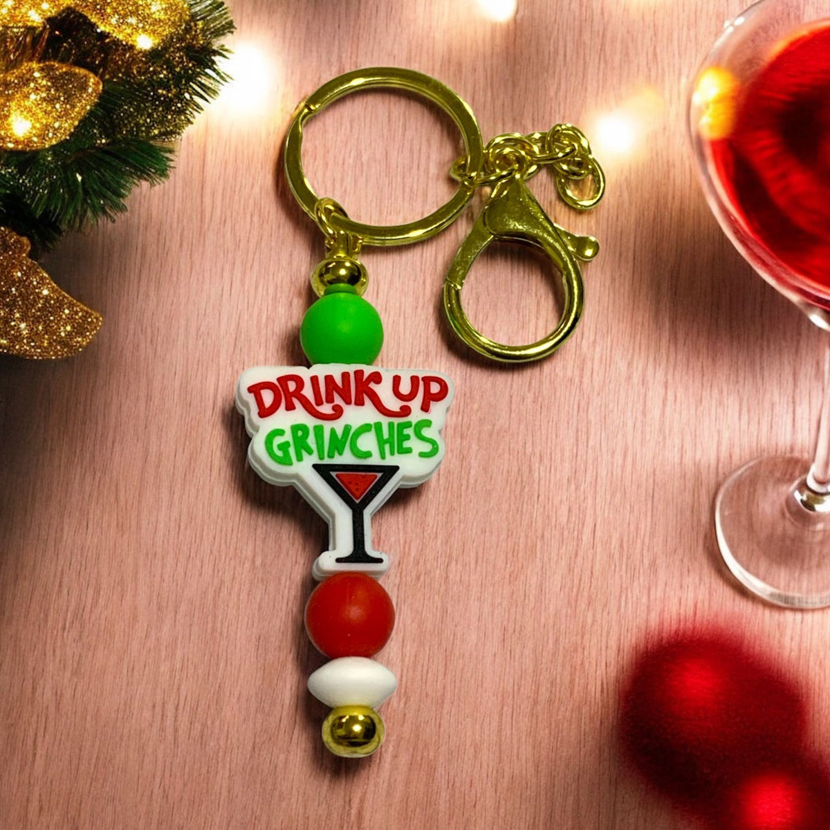 Keychain with 'Drink Up Grinches' text on a festive background with a glass of red wine and Christmas decorations.
