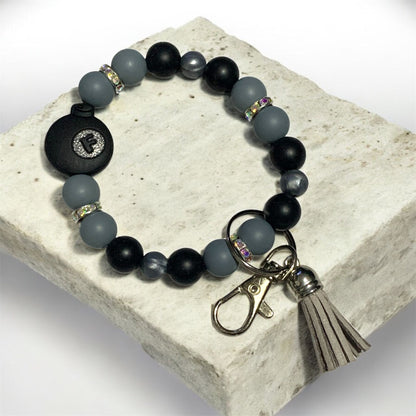 Bracelet with black and gray beads and a tassel charm on a stone surface. The focal bead is a black Bomb with the letter F.