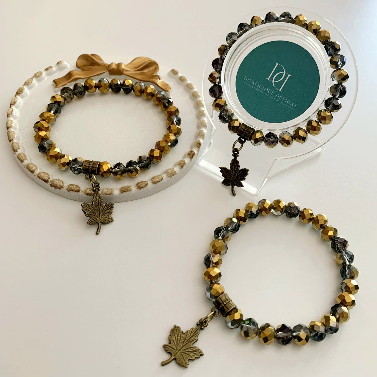 Two beaded golden chrome bracelets with leaf charms on a white surface, one on a stand with a brand label. Made by Divalicious Designs.