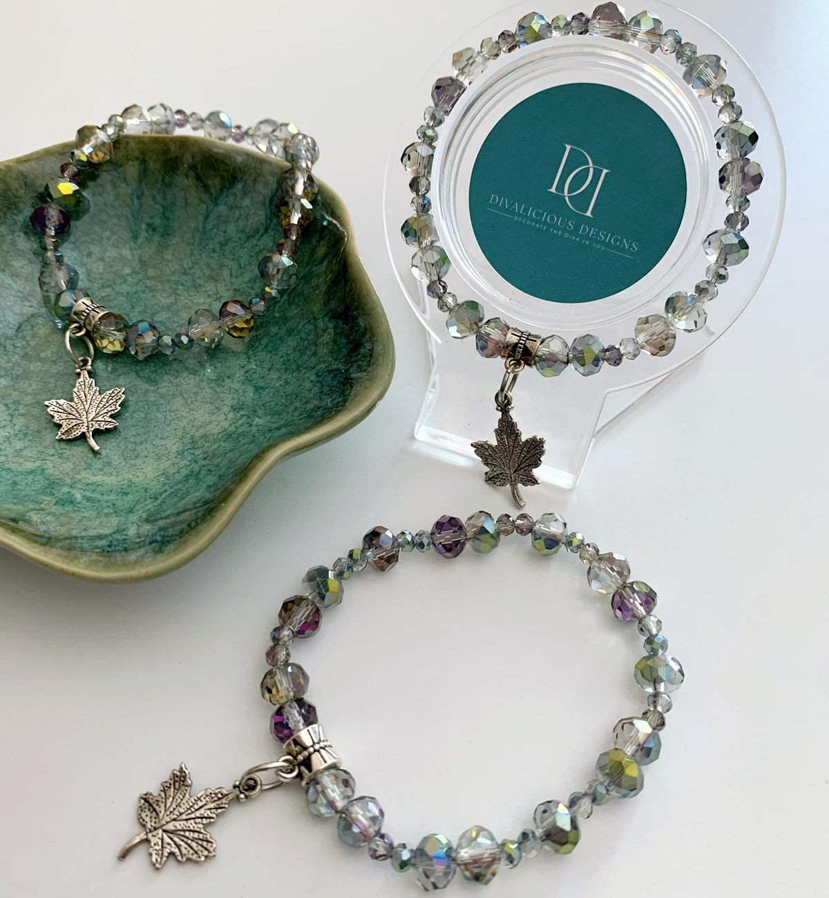 Silver and rainbow chrome beaded bracelet with leaf charm on a white surface, next to a green dish and a display stand. By Divalicious Designs.