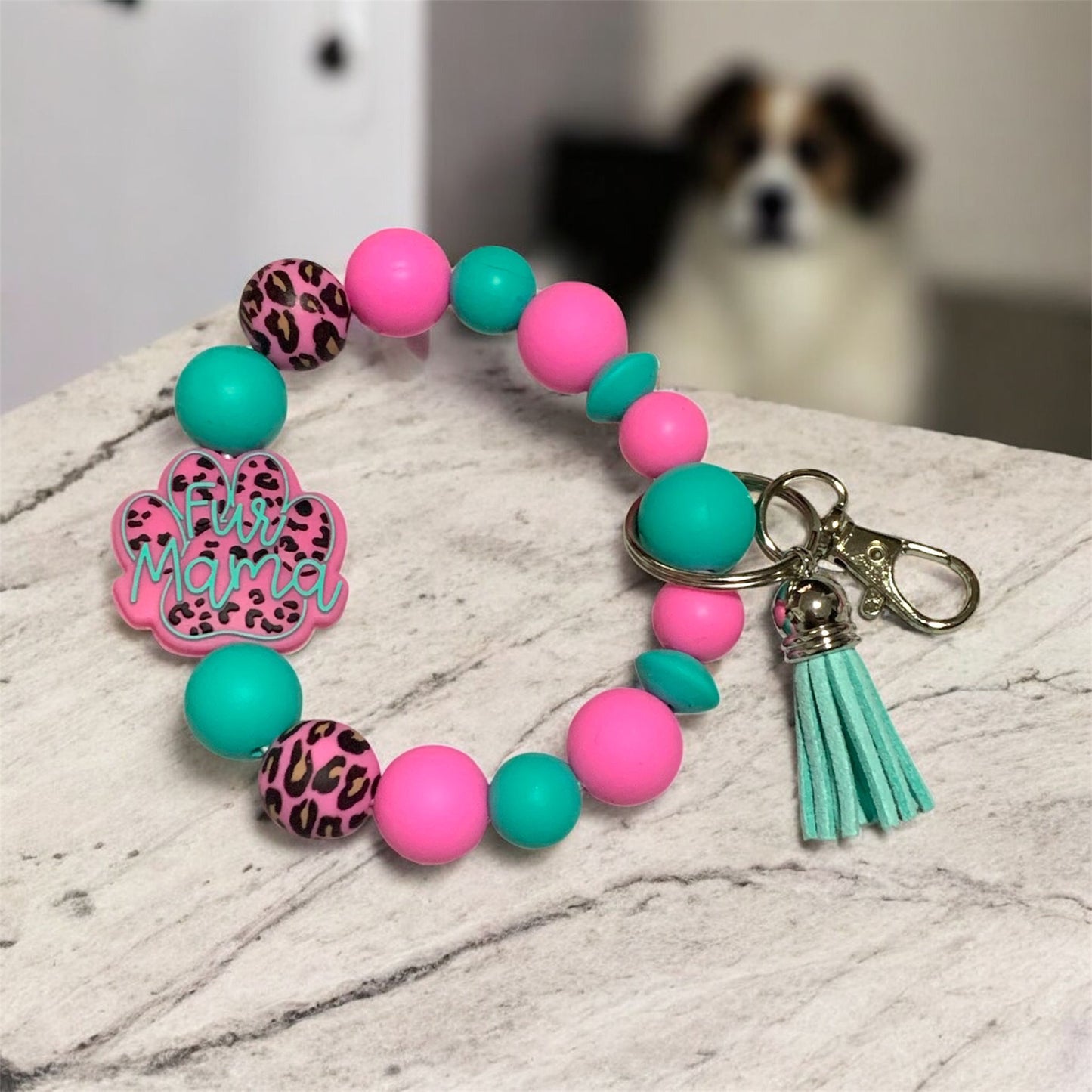 Teal and pink silicone keychain bracelet with a 'Fur Mama' focal bead,  displayed on a marble surface, with a dog in the background.