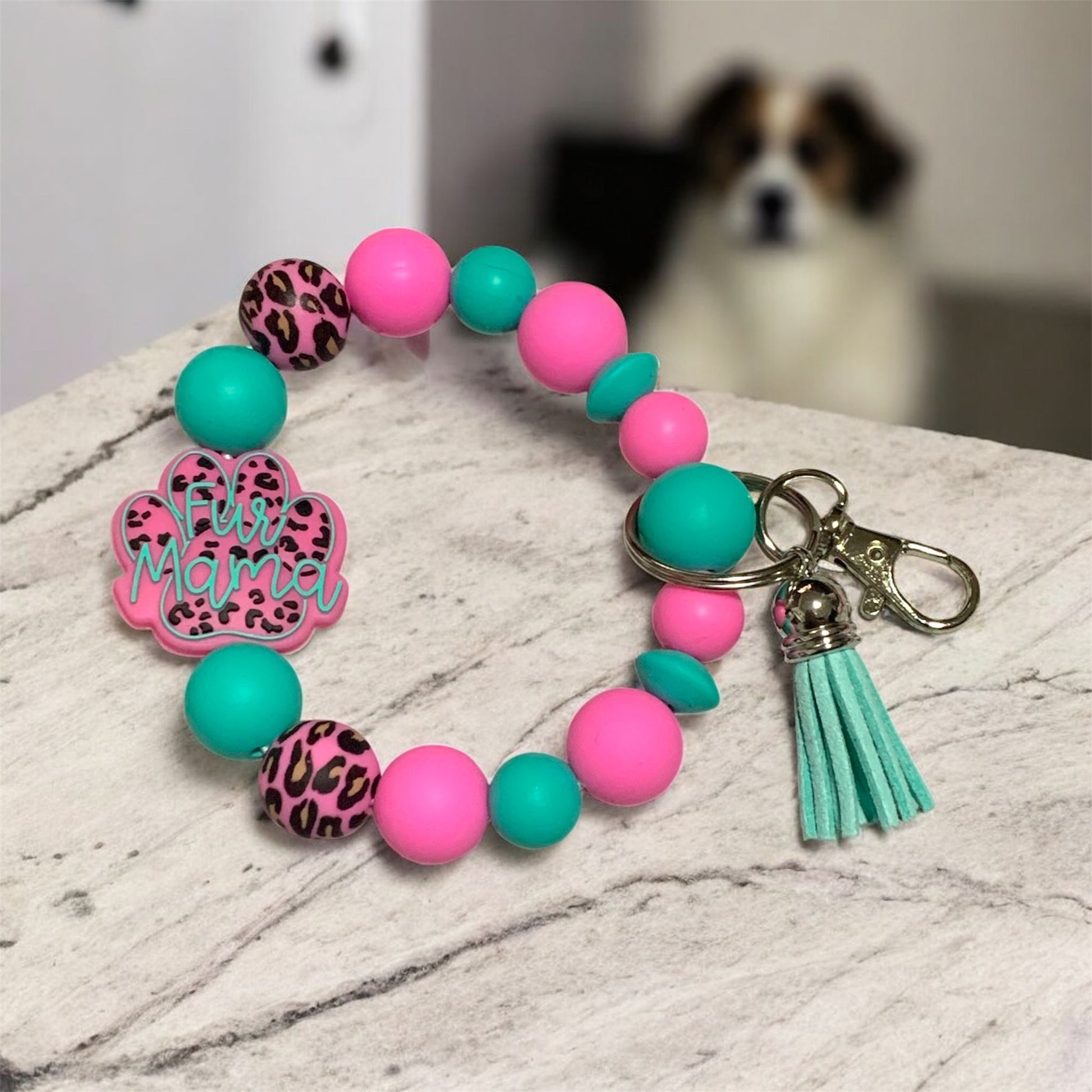 Teal and pink silicone keychain bracelet with a 'Fur Mama' focal bead,  displayed on a marble surface, with a dog in the background.