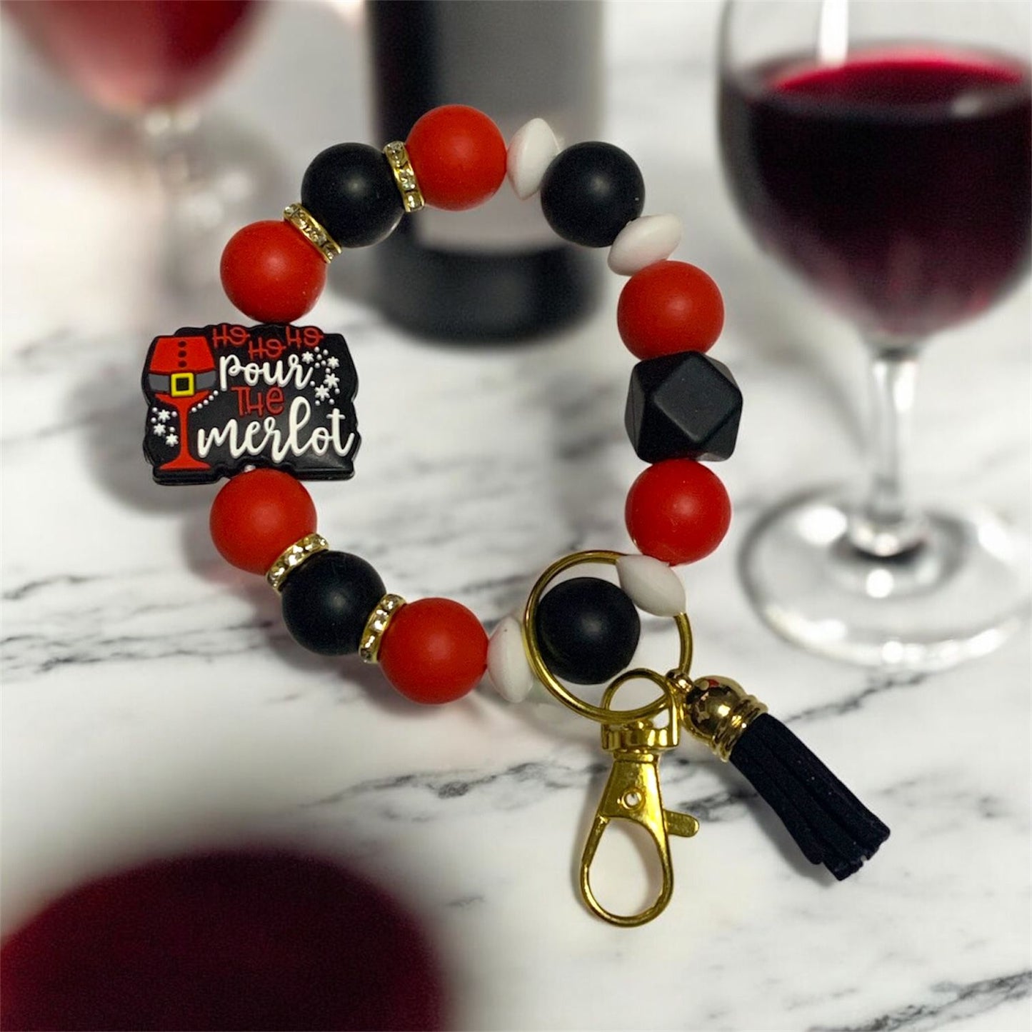 Bracelet with red, black, and white beads and a keychain on a marble surface with wine glasses in the background. The focal bead is a Santa inspired wineglass and the saying, "Ho Ho Ho, Pour The Merlot.".