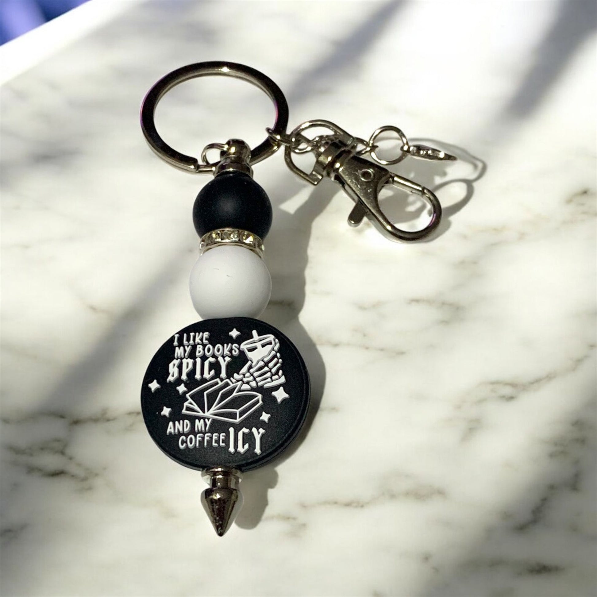 Keychain with a black and white focal bead that says, "I Like My Books Spicy, and My Coffee Icy,"  on a marble surface
