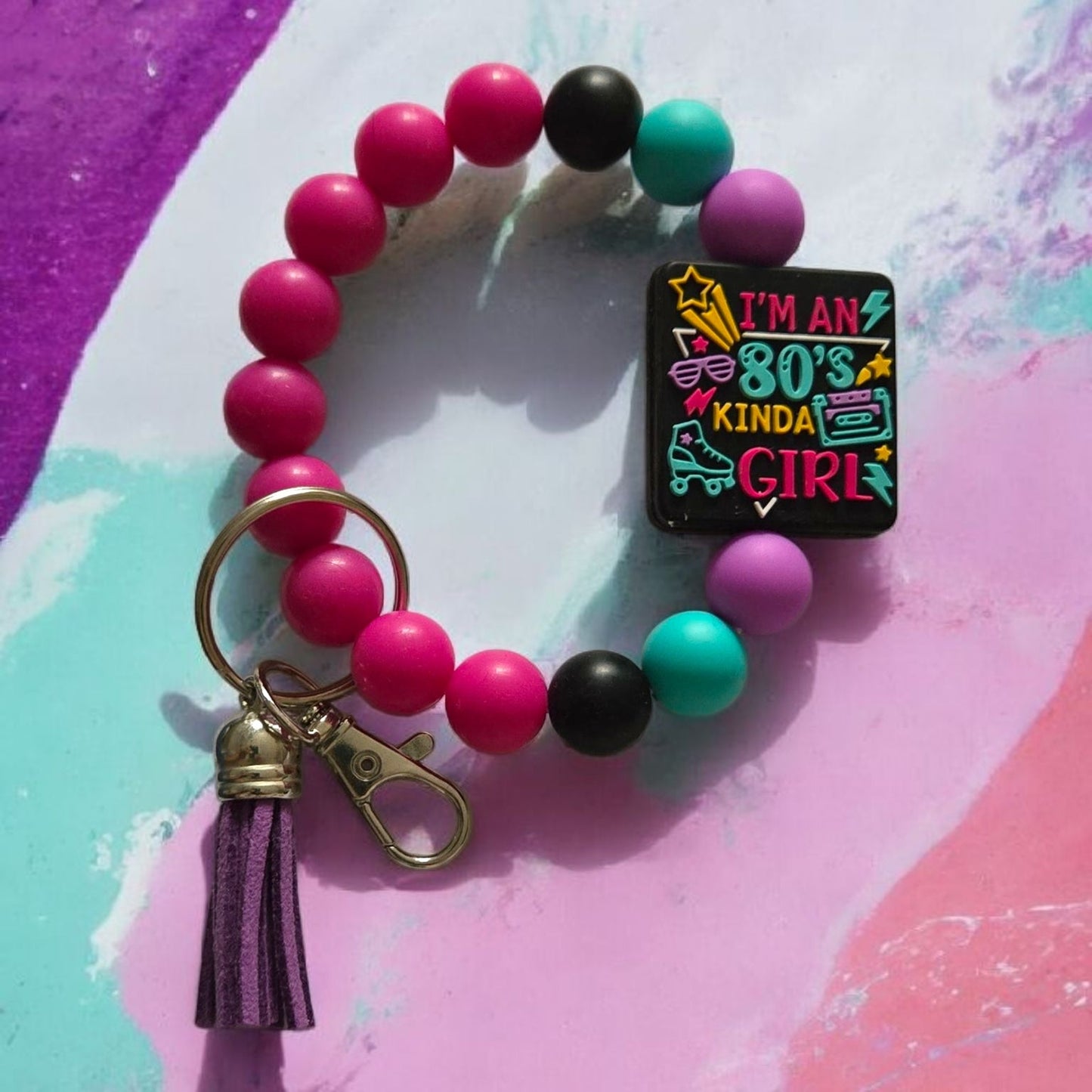 Colorful beaded bracelet with the focal bead saying, "I'm an 80's Kinda Girl" with a keychain and tassel on a multicolored background
