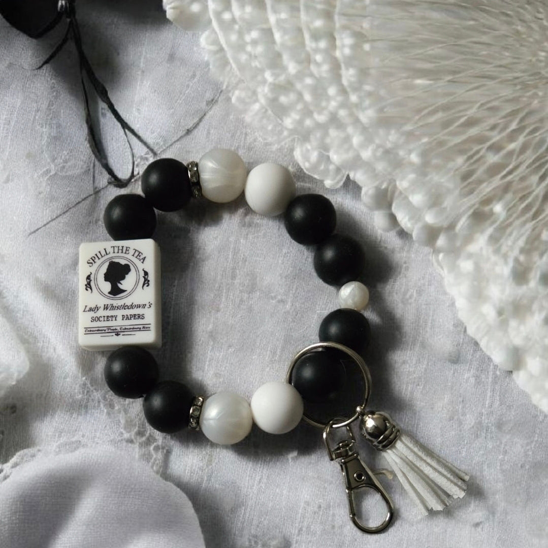 Lady Whistledown black and white beaded keychain wristlet with a Spill The Tea focal bead, on a textured surface, inspired by Lady Whistledown and Bridgerton.
