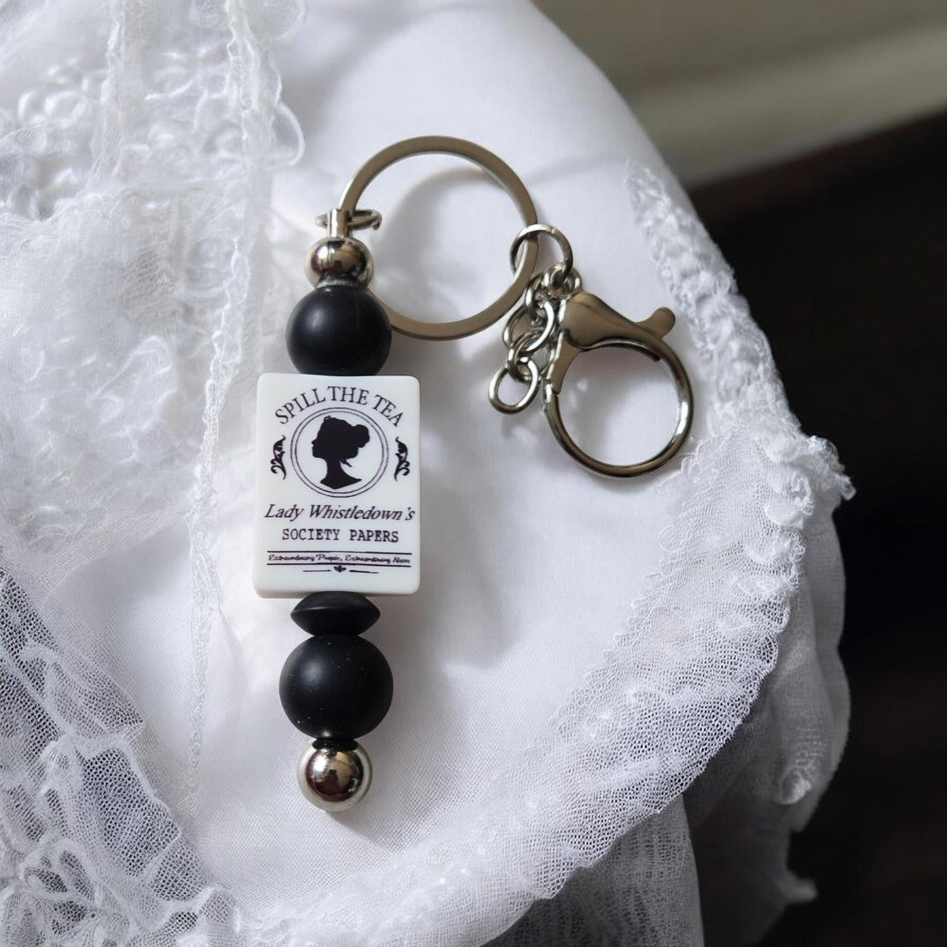 Lady Whistledown, Spill the Tea Keychain with black and silver beads on a white lace background. Inspired by Bridgerton.
