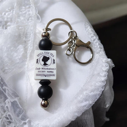 Lady Whistledown, Spill the Tea Keychain with black and silver beads on a white lace background. Inspired by Bridgerton.