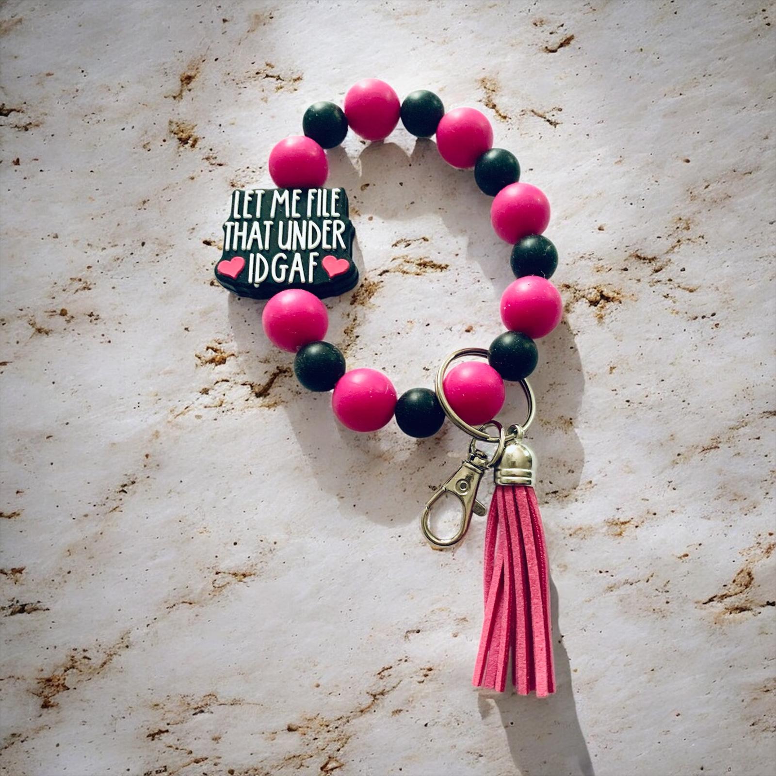 Bracelet with pink and black beads and a tassel on a marble surface, the focal bead states, "Let Me File That Under IDGAF."