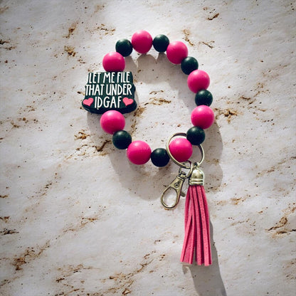 Bracelet with pink and black beads and a tassel on a marble surface, the focal bead states, "Let Me File That Under IDGAF."
