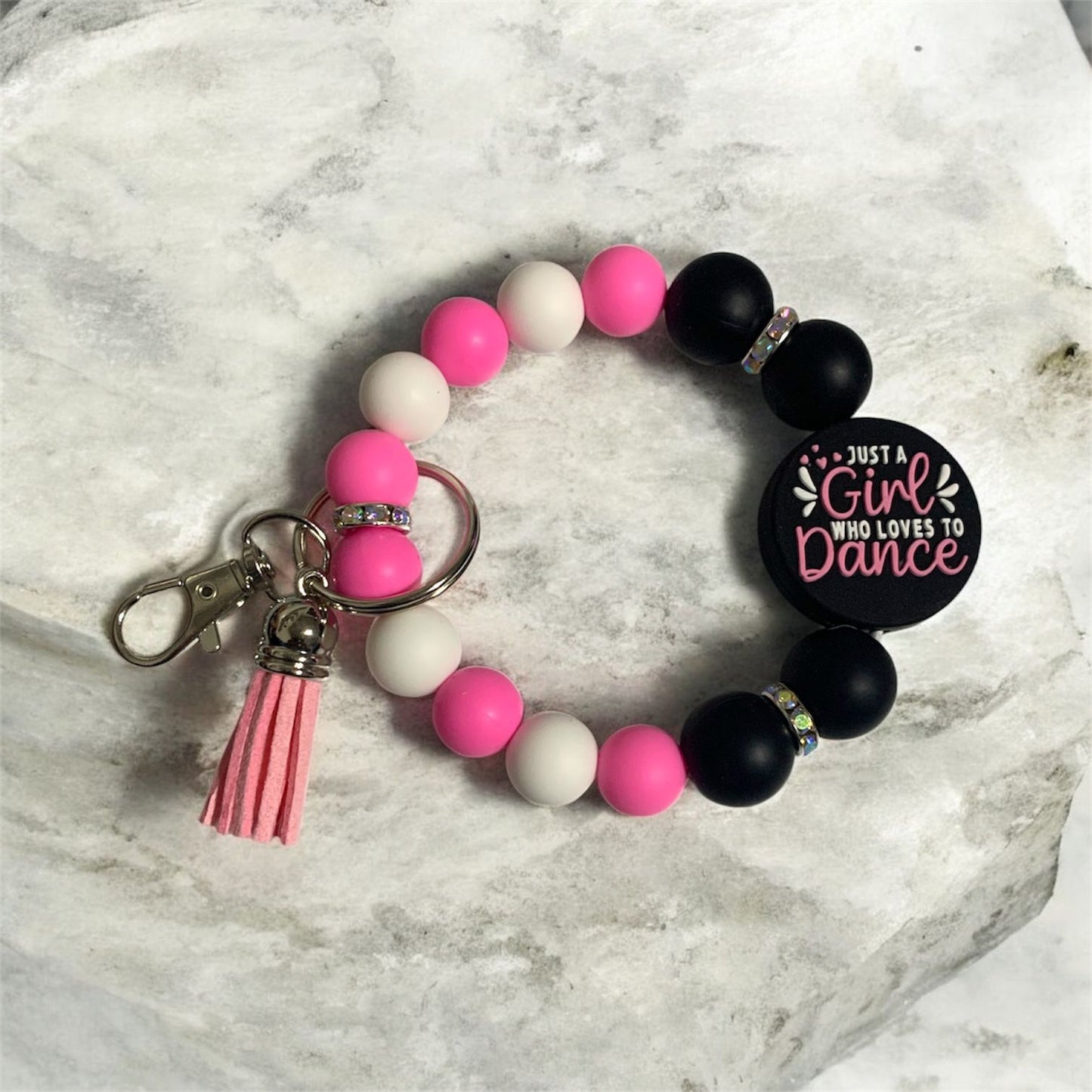 Beaded bracelet with pink, white, and black beads, a tassel, and a keychain on a marble surface. The focal bead says Just a Girl Who Loves to Dance.