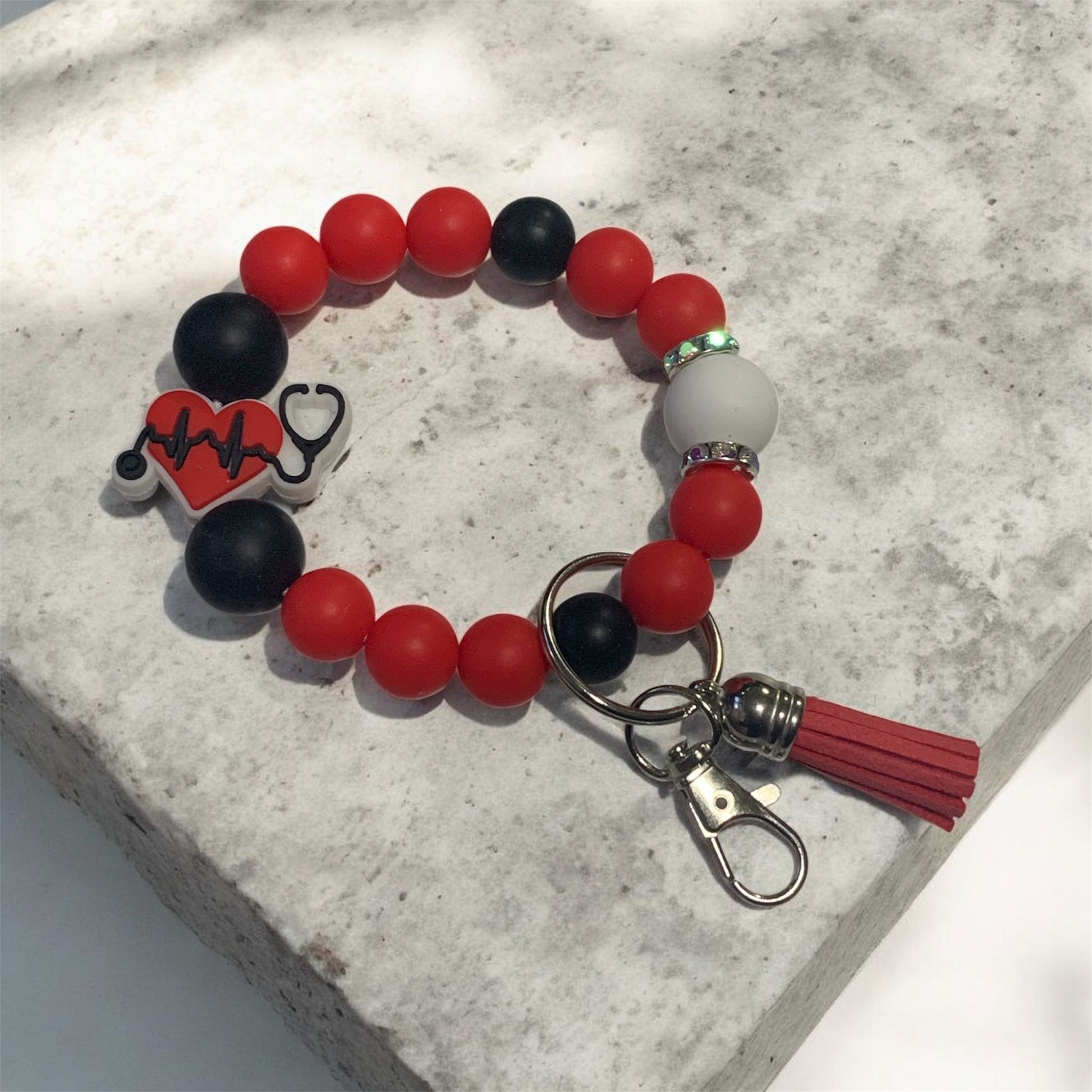 Keychain Bracelet with red, black, and white beads on a textured surface. The focal bead is Stethoscope with a lifeline and heart.