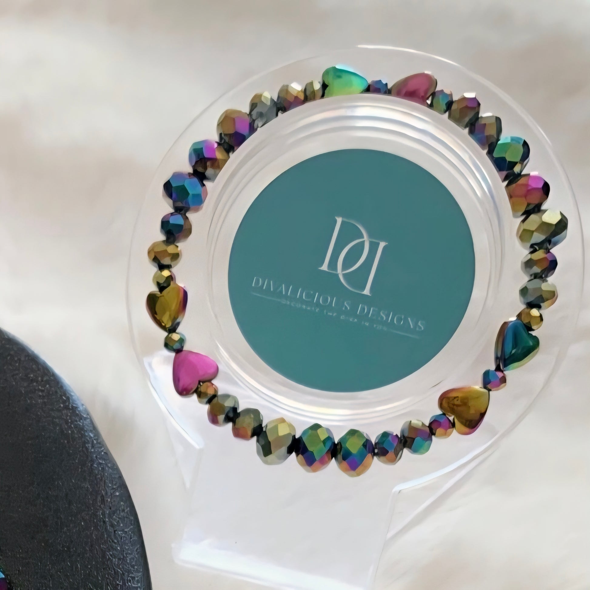 A Multi-chrome Crystal Bracelet with hearts hung on clear holder with Divalicious Designs logo which is resting on a light marble surface.  , Photo and Bracelet made by Divalicious Designs.