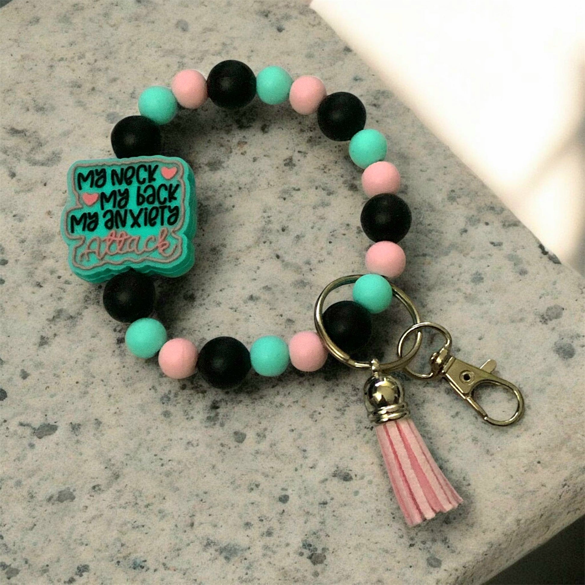 Colorful beaded bracelet with a keychain and tassel on a gray surface, The focal bead states, My Neck, My Back, My Anxiety Attack.