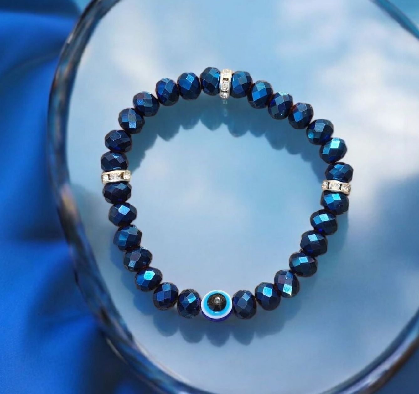 Navy chrome crystal bracelet with rhinestone accents and a centered evil eye protection bead displayed on a blue reflective surface.