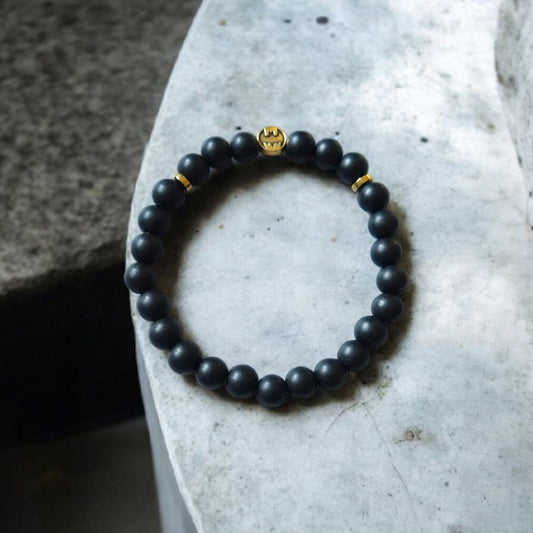 Matte onyx beaded bracelet with gold Batman-style charm displayed on a stone slab.