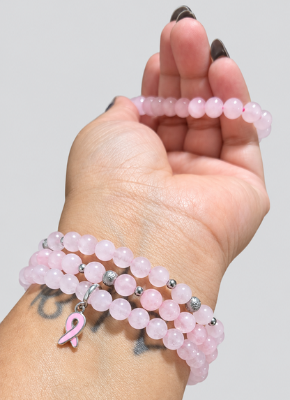 A close-up of a person's wrist wearing three layered pink beaded bracelets, including a small pink breast cancer awareness ribbon charm, against a plain light gray background.