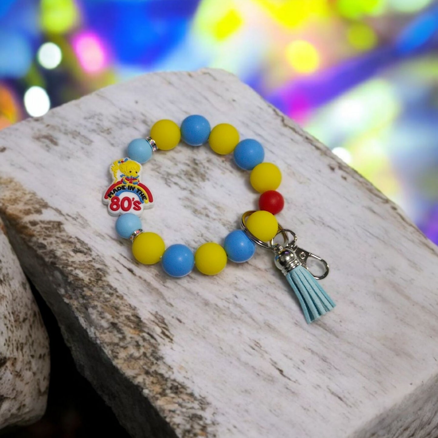 Colorful beaded bracelet with a tassel charm on a wooden surface with a blurred colorful background. The focal bead is Rainbow Brite on a Rainbow saying, "Made in the 80'S".