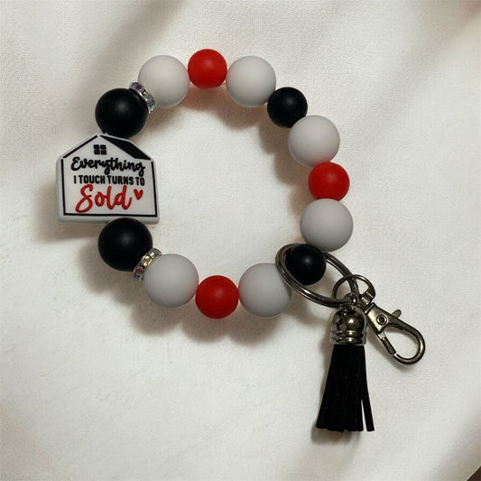 Keychain bracelet with a real estate them off a house focal bead stating, " everything I touch turns to sold". Item is displayed on a light counter