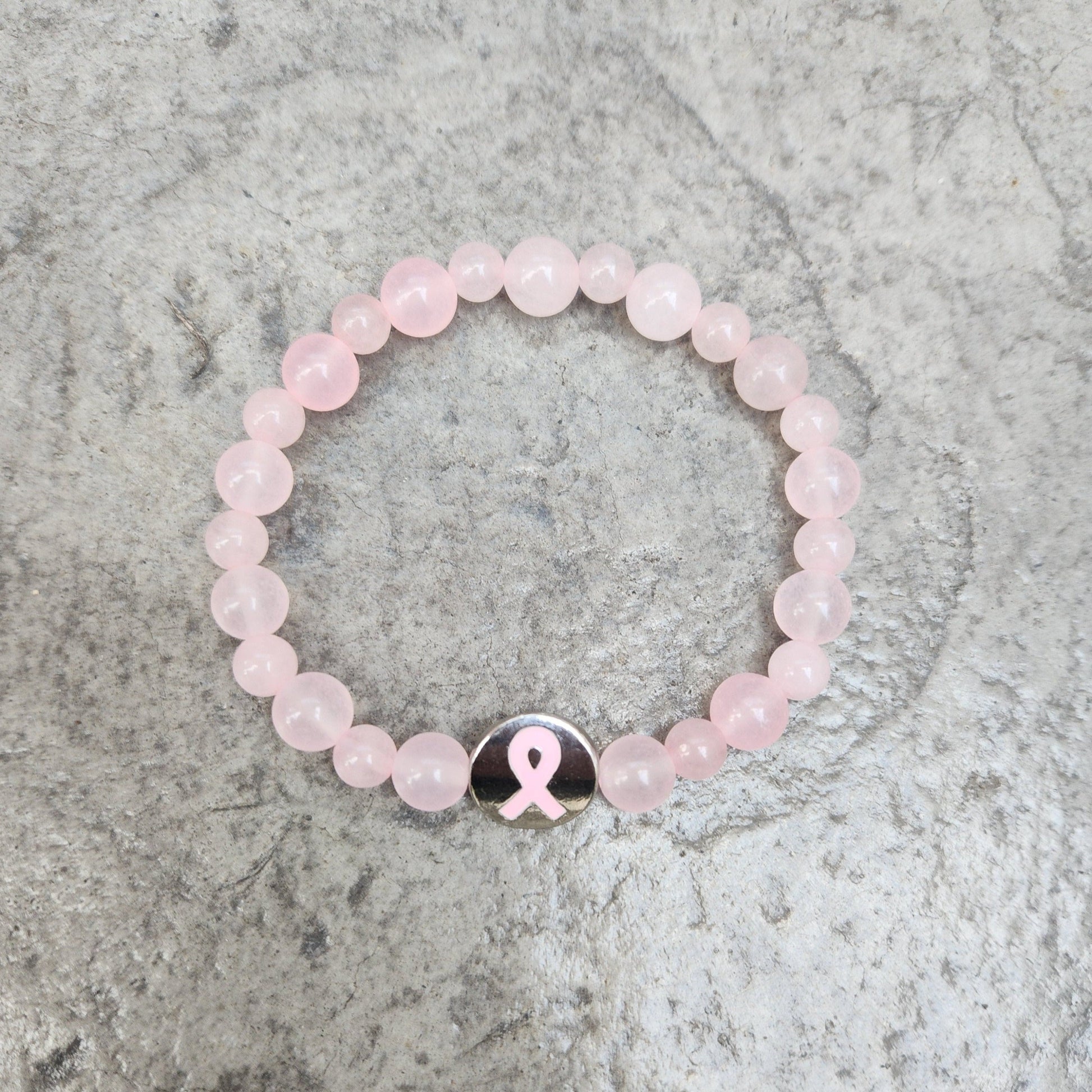 A bracelet made of rose quartz beads with a metal breast cancer awareness charm as the centerpiece, displayed against a textured background.