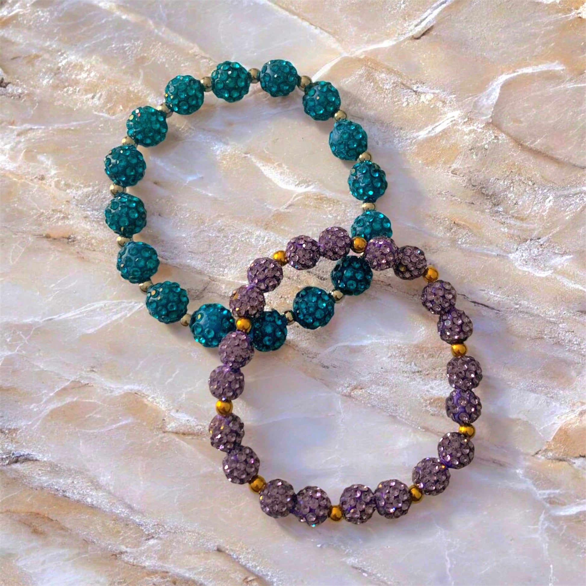 Two Shamballa and stainless Steel beaded bracelets, one teal and one lilac, on a textured surface.
Photo and bracelets made by Divalicious Designs.