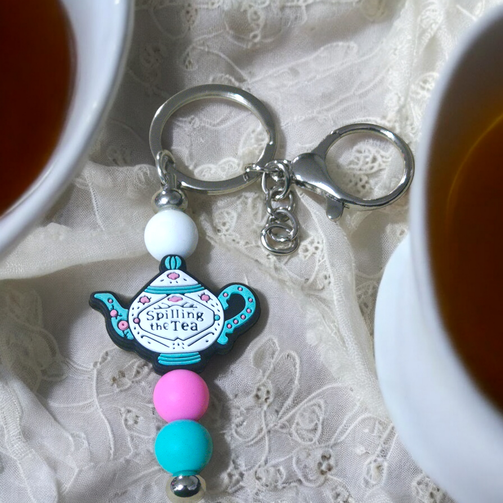 Keychain with teapot design thar says,  Spilling The Tea, on a lace background