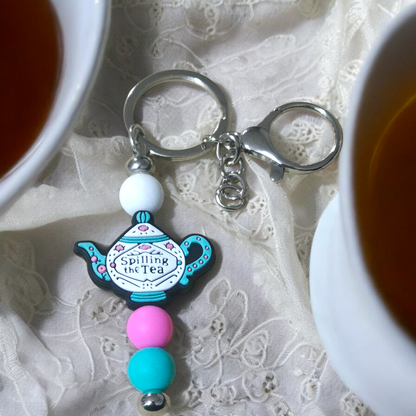 Keychain with teapot design thar says,  Spilling The Tea, on a lace background
