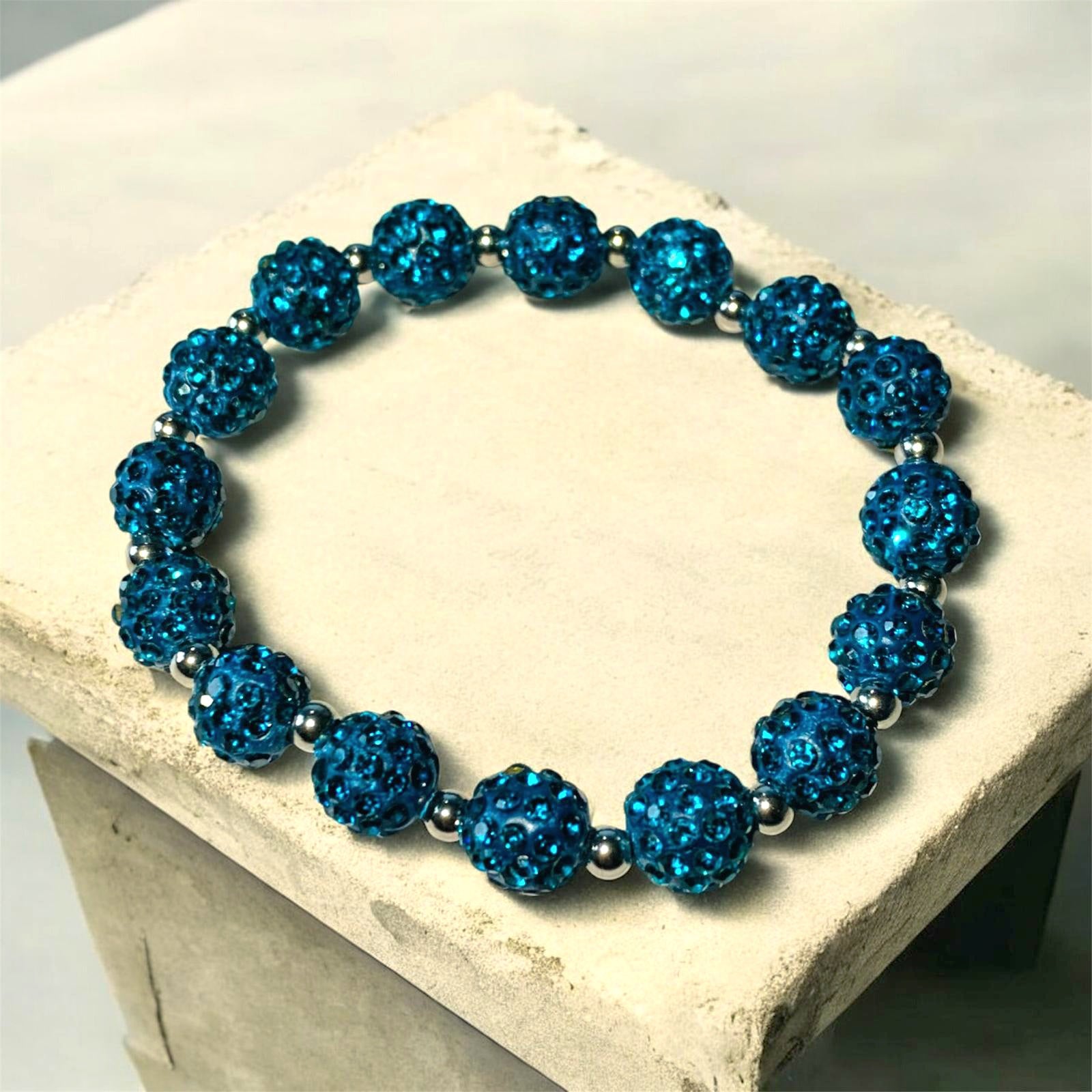 A Bracelet made of Teal Shamballa beads and stainless steel beads laying on a light stone surface. Picture and bracelet by Divalicious Designs.