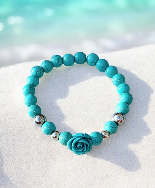 Turquoise beaded bracelet with stainless steel accents and a reconstituted turquoise rose focal bead displayed on a white sand-like surface.