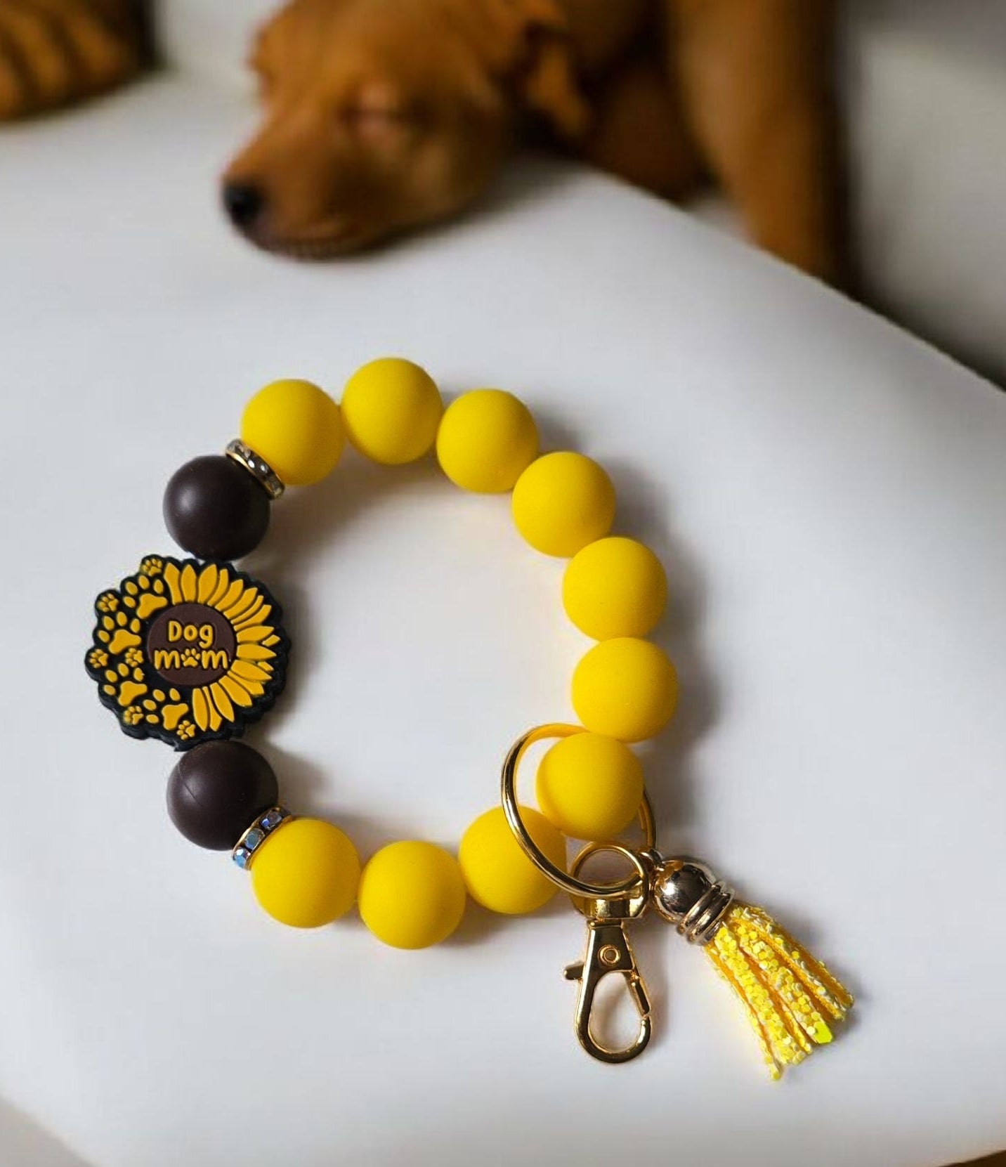 Yellow beaded silicone keychain bracelet with a tassel and focal bead of a sunflower with the wirds, Dog Mom. It is displayed on a white surface, with a dog in the background.